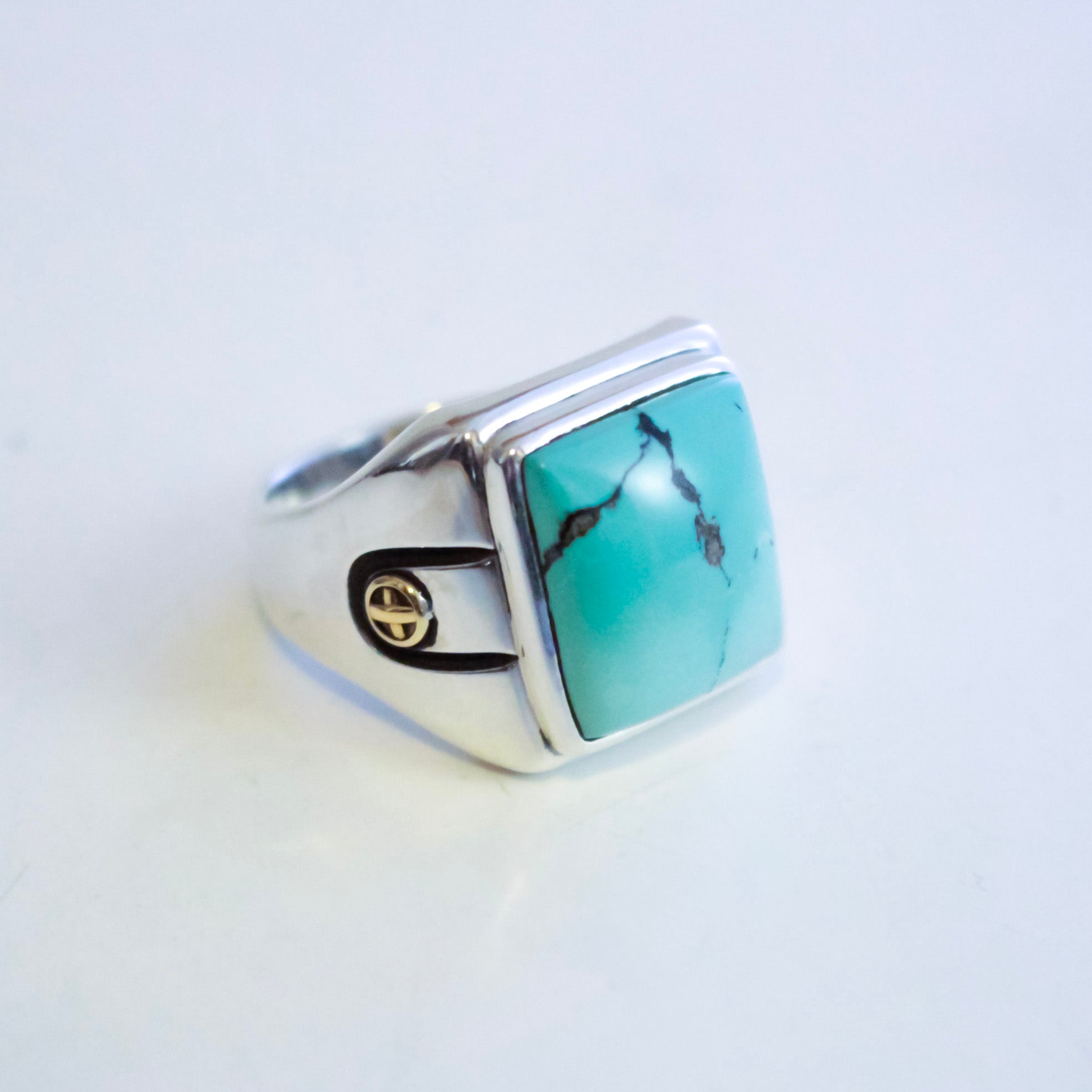 A sterling silver ring featuring a square hubei turquoise stone with natural dark veining, accented by 2 small gold emblems on the band, set against a plain white background.