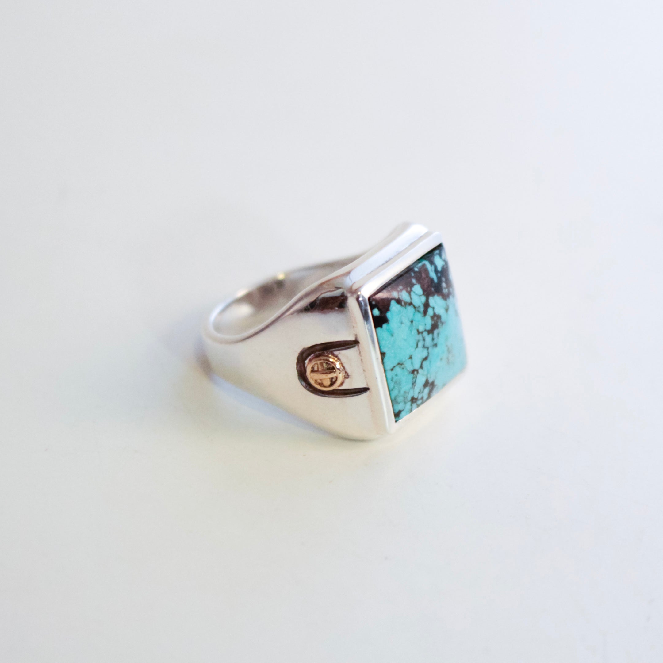 A Sterling Silver signet ring featuring a square Hubei turquoise stone with dark matrix, accented by a small 18K Gold bosses on the band, set against a plain white background.