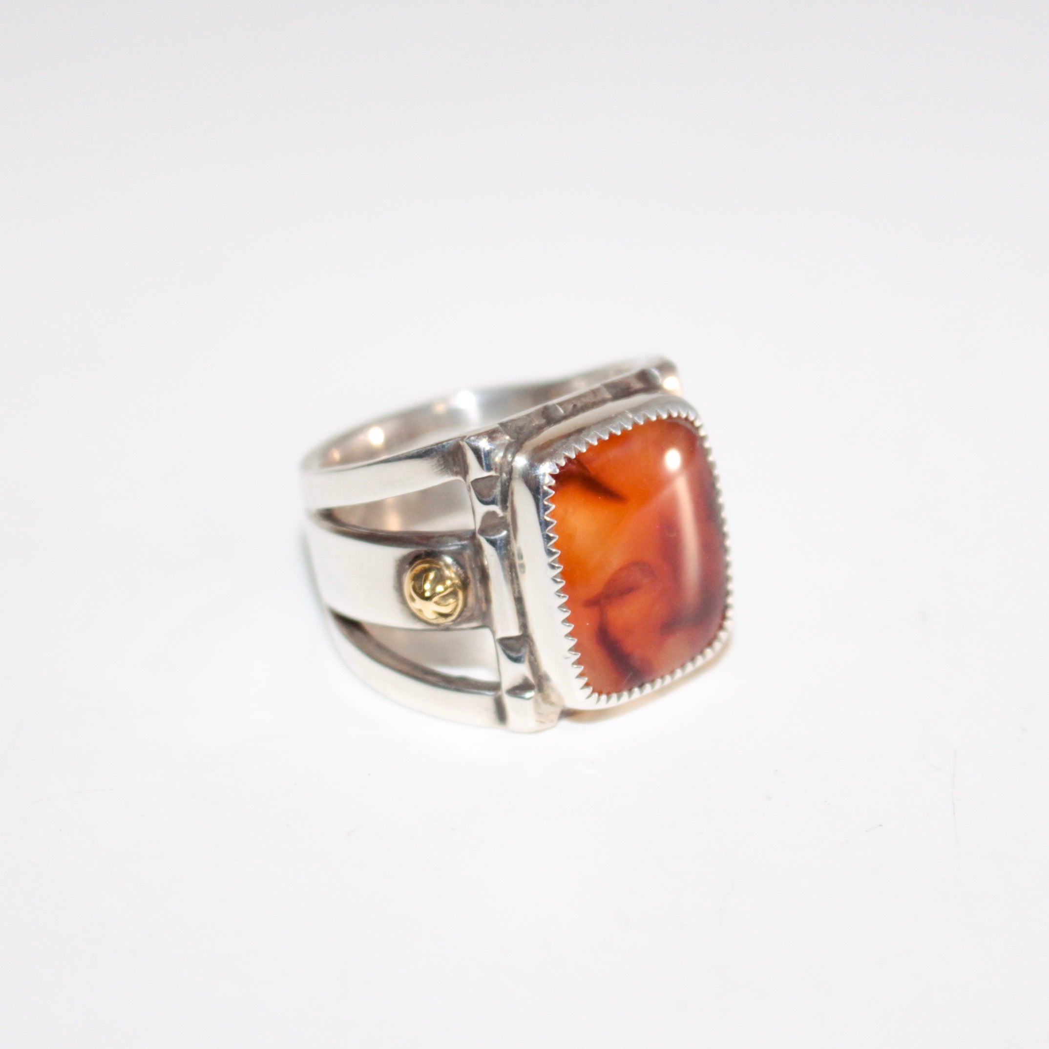 A silver ring featuring a large, square, amber-orange marbled Cherry Carnelian in a bezel setting, with small 22K gold accent bosses on the band, shown on a plain white background.