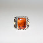 A silver ring featuring a large, square, amber-orange marbled Cherry Carnelian in a bezel setting, with  small 22K gold accent bosses on the band, shown on a plain white background.