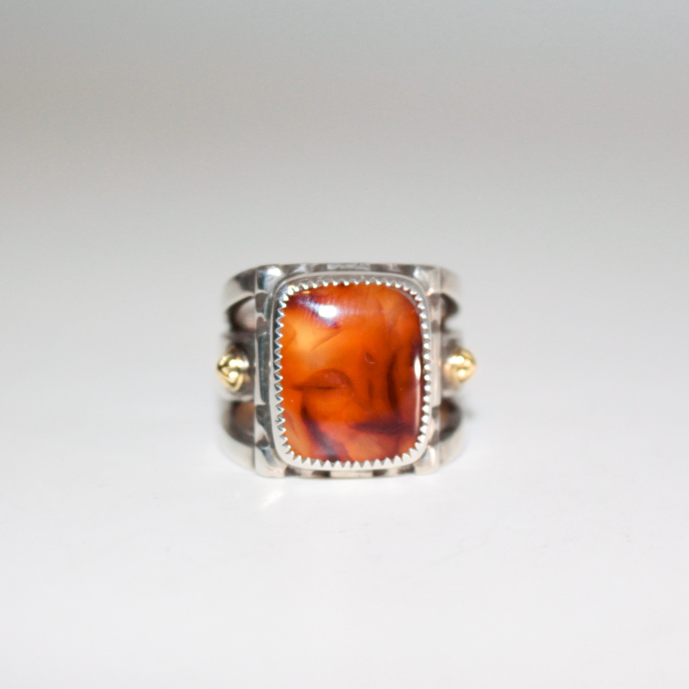 A silver ring featuring a large, square, amber-orange marbled Cherry Carnelian in a bezel setting, with  small 22K gold accent bosses on the band, shown on a plain white background.