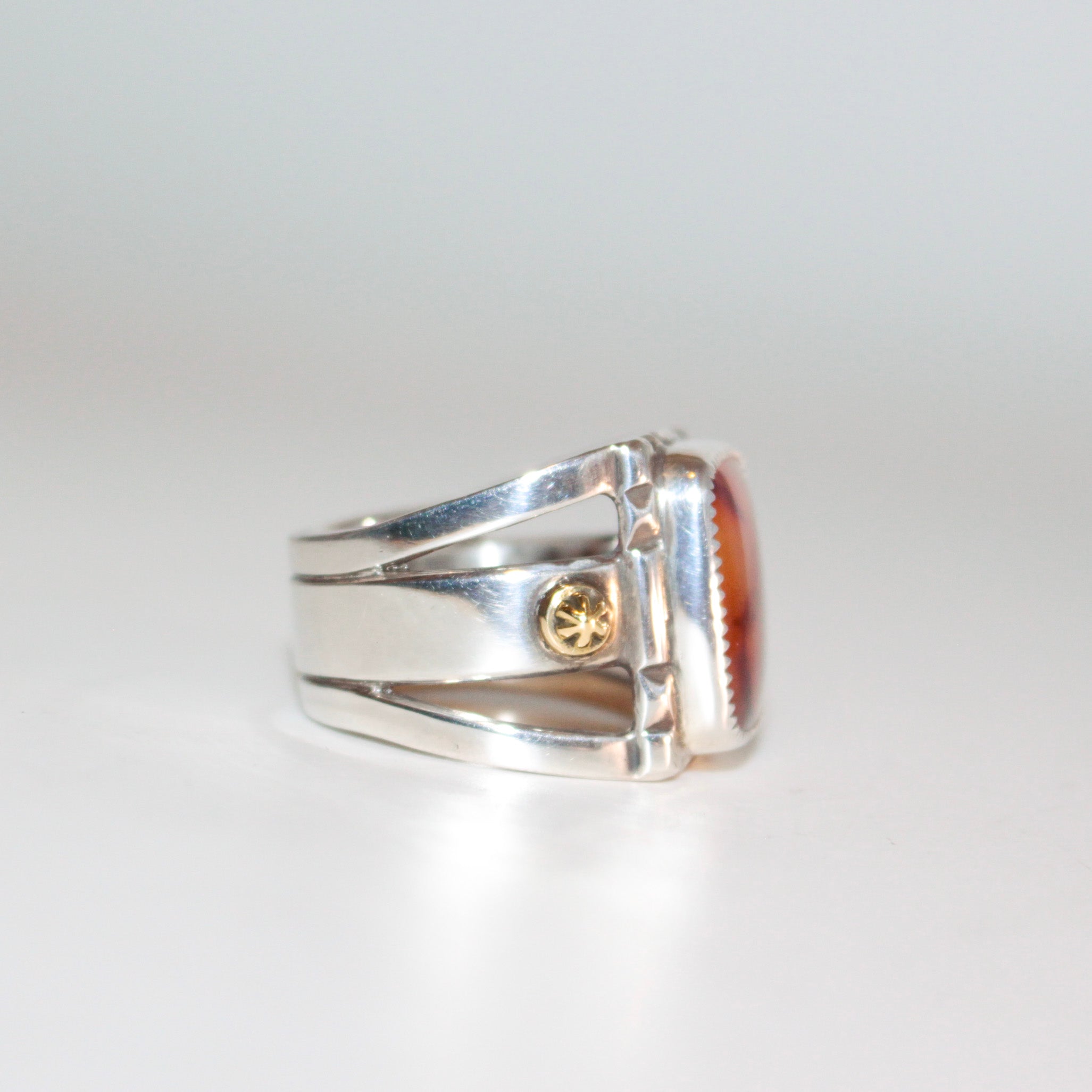 A silver ring featuring a large, square, amber-orange marbled Cherry Carnelian in a bezel setting, with small 22K gold accent bosses on the band, shown on a plain white background.