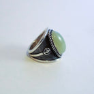 A silver-toned ring featuring a large, smooth oval light yellow jade in a bezel setting, with textured dark silver sterling silver bands and a small circular detail on the side, set against a plain light background.
