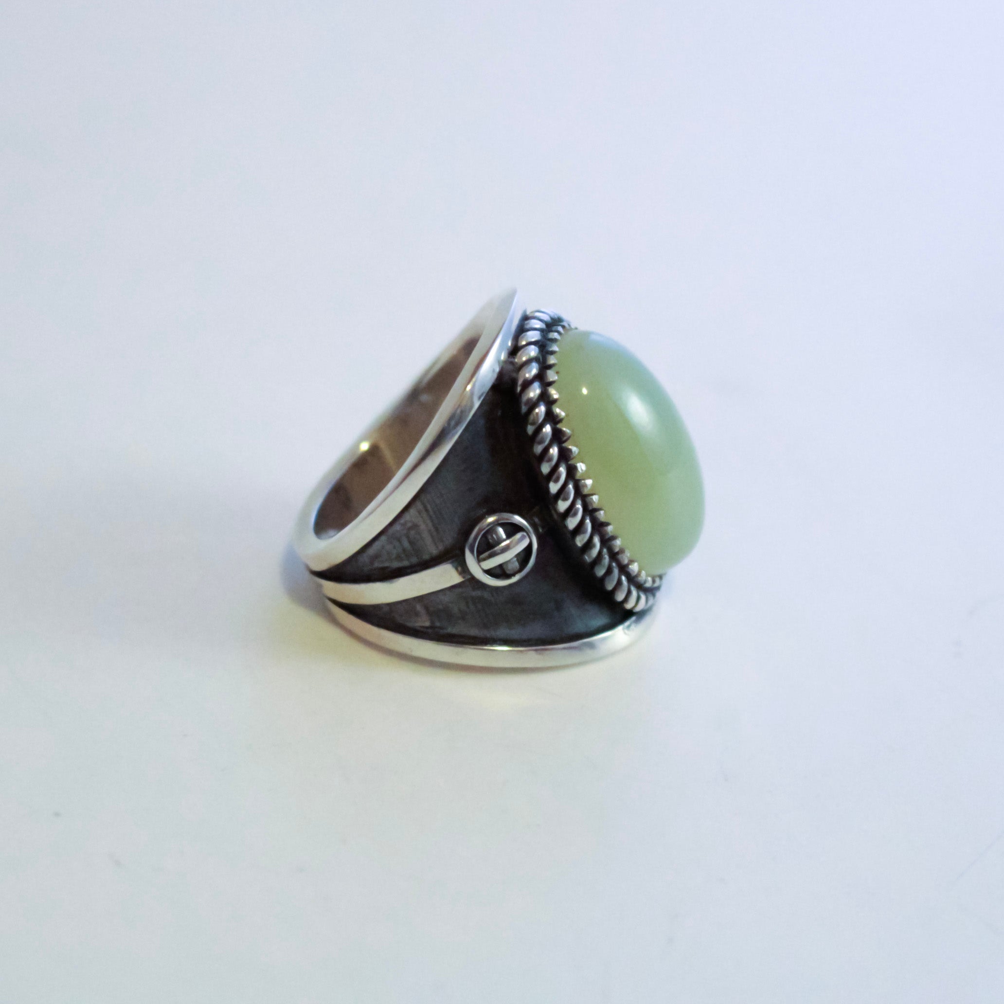 A silver-toned ring featuring a large, smooth oval light yellow jade in a bezel setting, with textured dark silver sterling silver bands and a small circular detail on the side, set against a plain light background.