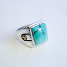 A sterling silver ring featuring a square hubei turquoise stone with natural dark veining, accented by 2 small gold emblems on the band, set against a plain white background.