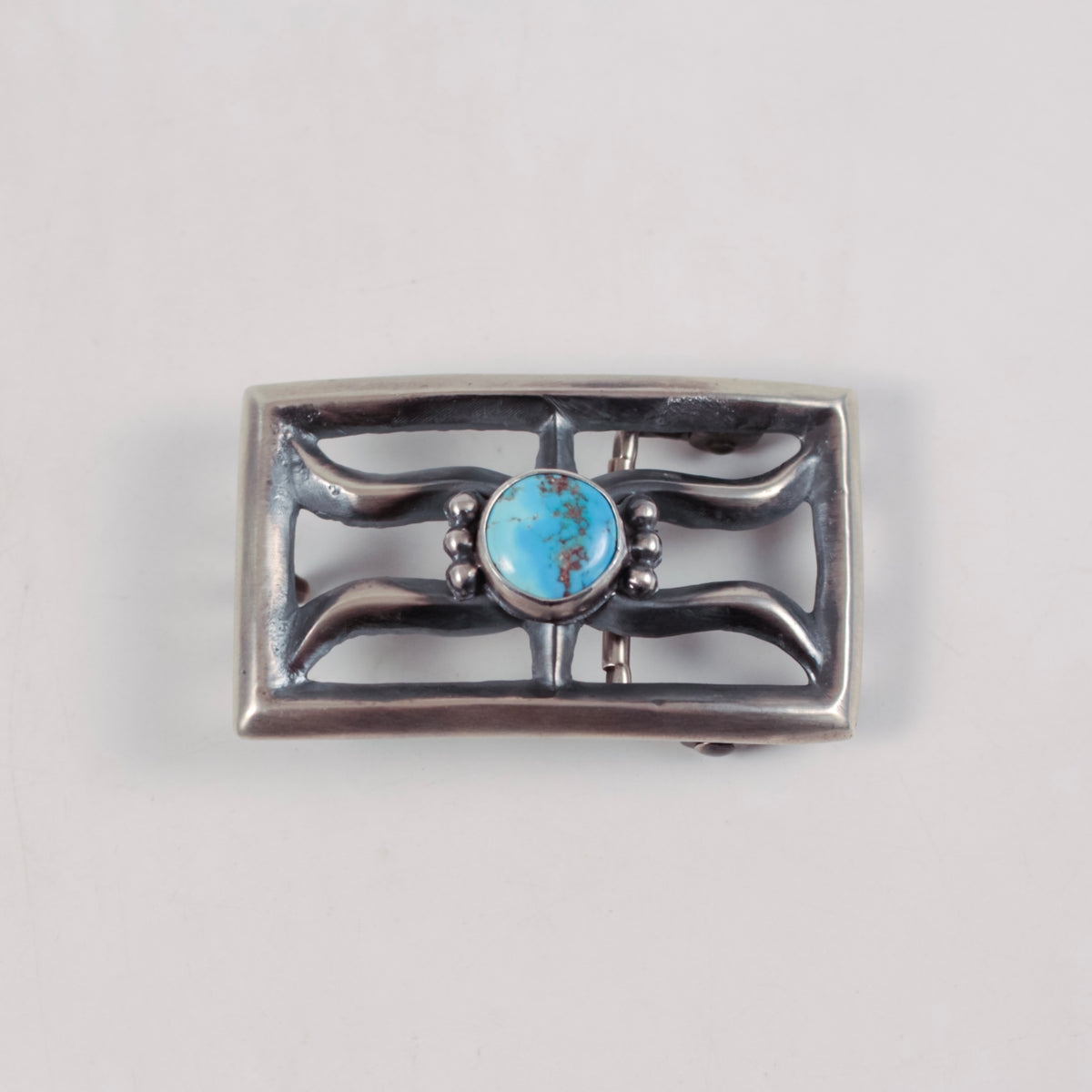 A rectangular sterling silver belt buckle featuring a central oval turquoise stone surrounded by decorative curved silver bars and small bead accents, shown on a plain light gray background.