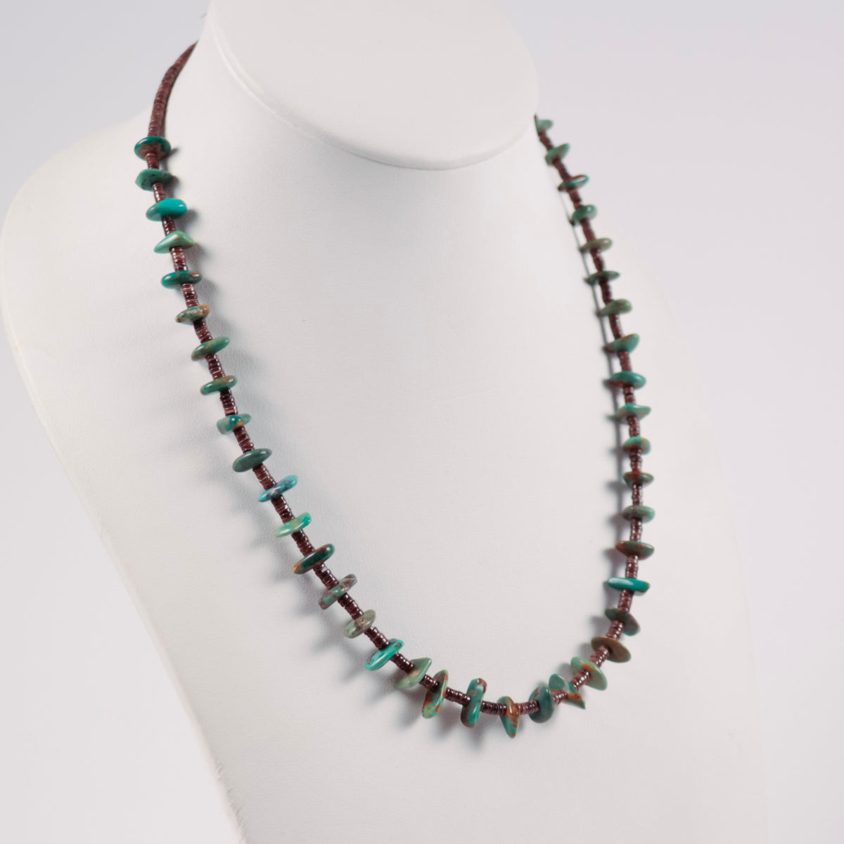 A beaded necklace displayed on a white mannequin bust. The necklace features small, irregular turquoise stone chips spaced by smaller pin shell beads. 