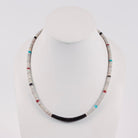 A beaded necklace displayed on a white mannequin bust against a light gray studio background. The necklace features predominantly off-white and gray cylindrical beads, with a dark charcoal/black curved section at the bottom center and small accent beads in turquoise blue, red, and black.