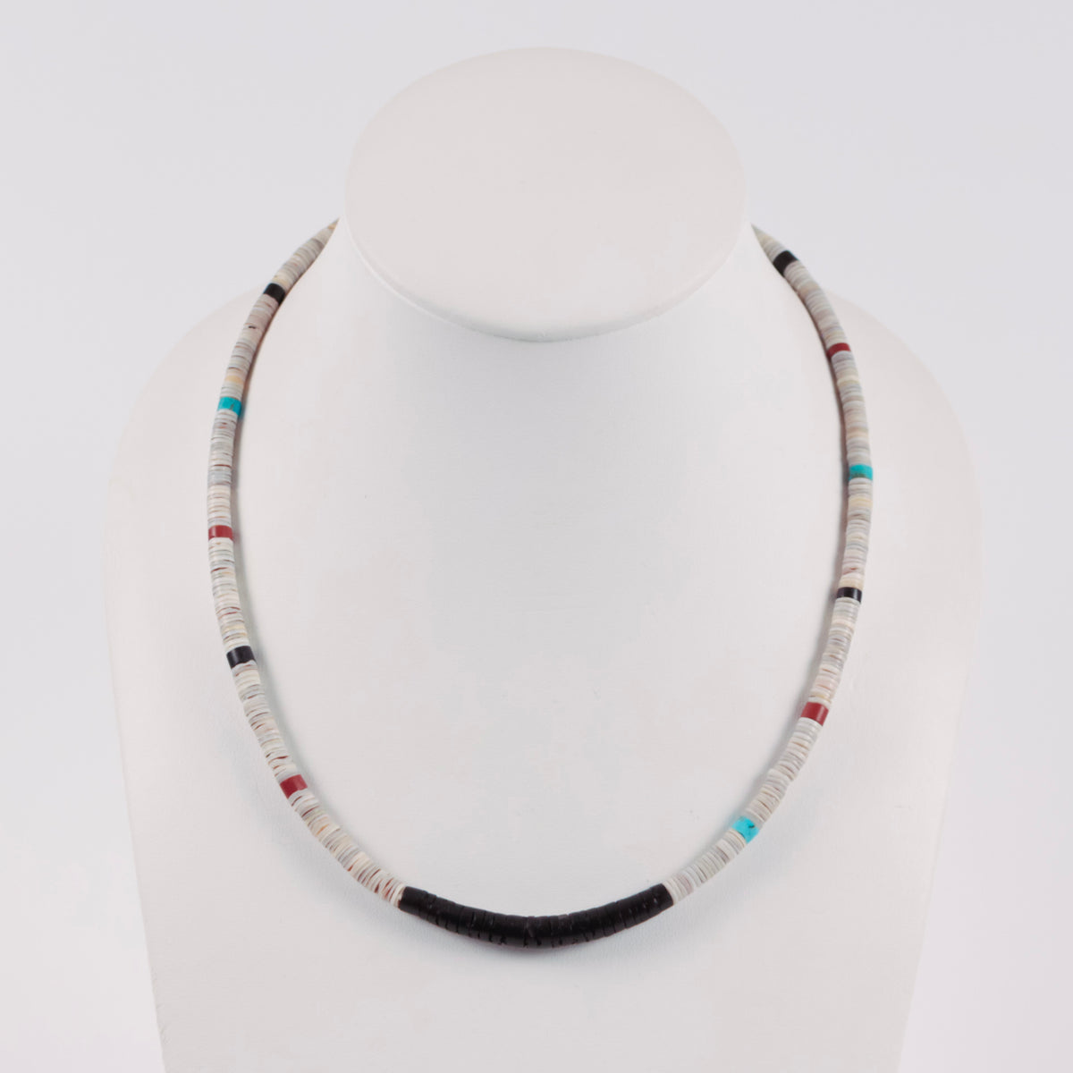 A beaded necklace displayed on a white mannequin bust against a light gray studio background. The necklace features predominantly off-white and gray cylindrical beads, with a dark charcoal/black curved section at the bottom center and small accent beads in turquoise blue, red, and black.